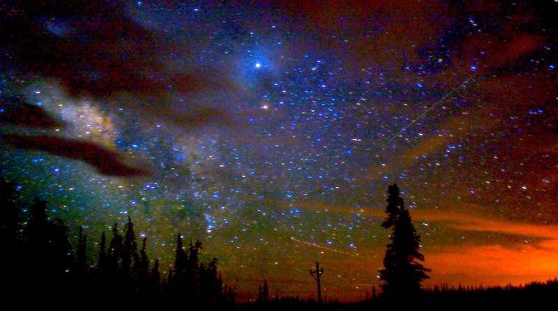 An image of a night sky in a rural area. The sky is star filled, with some clouds, and hues of blue and orange. There are shadows of trees in the foreground as well as evidence of human habitation in the form of a utility pole.