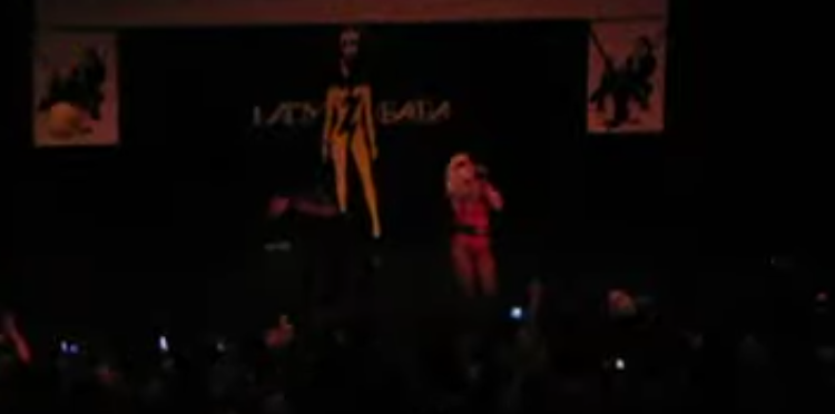 Photo of Lady Gaga and her dancer performing on stage. The venue is darkened. She is wearing a homemade red outfit.