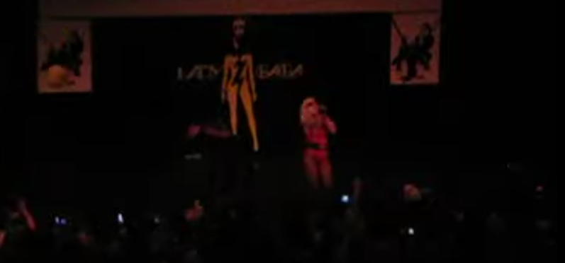 Photo of Lady Gaga and her dancer performing on stage. The venue is darkened. She is wearing a homemade red outfit.