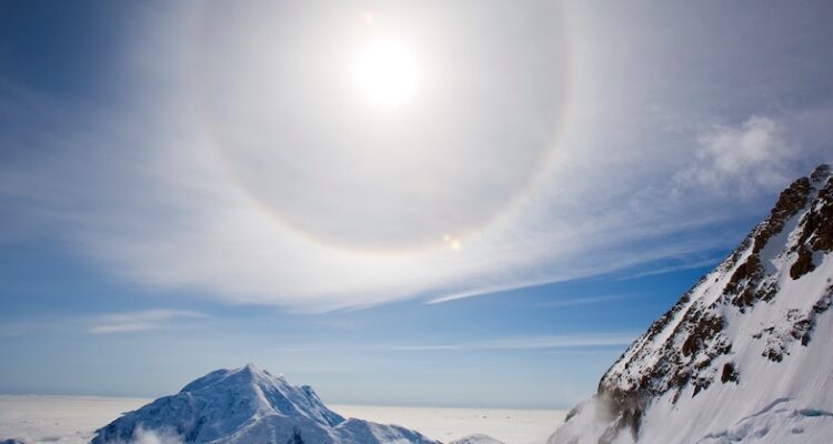 A halo around the sun, over a snowy landscape, with mountain peaks in the foreground. The sky is party cloudy but bright.