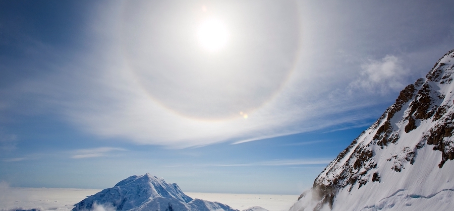 A halo around the sun, over a snowy landscape, with mountain peaks in the foreground. The sky is party cloudy but bright.
