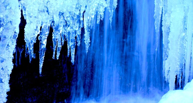 Icicles in the foreground with a waterfall in the background. The ice and water have a blue tint to them.