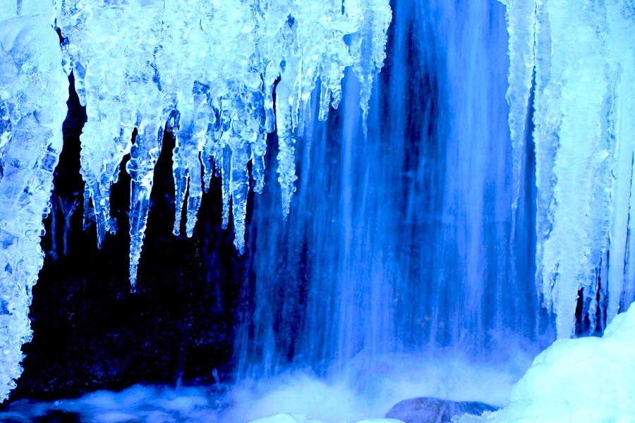 Icicles in the foreground with a waterfall in the background. The ice and water have a blue tint to them.