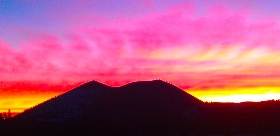 The outline of a backlit mountain. In the background is a colorful sky, illuminated by bright pink, yellow and purple clouds at sunset.
