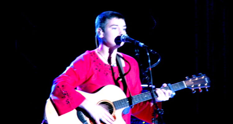 A person, Sinéad O'Connor, on a stage with a boom microphone, holding a guitar. SHe is wearing a red outfit, and the background is dark.