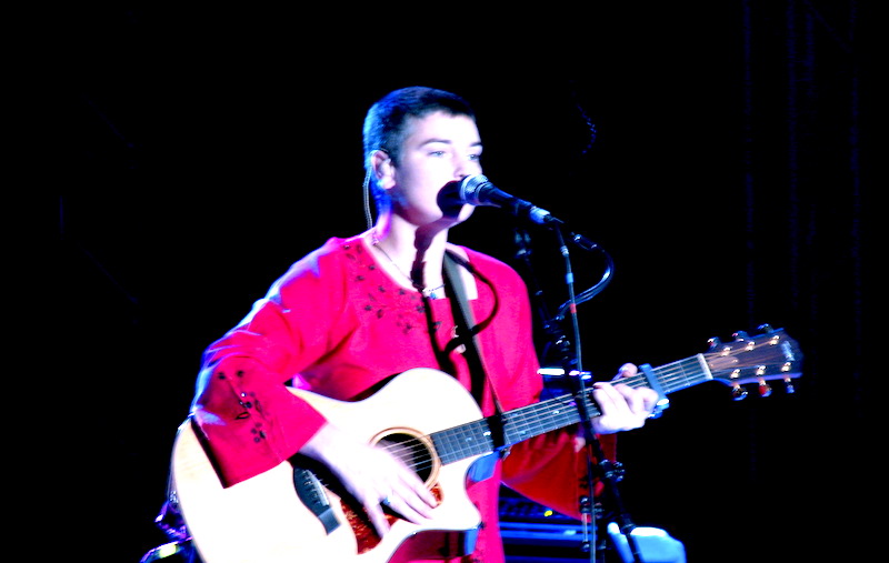A person, Sinéad O'Connor, on a stage with a boom microphone, holding a guitar. SHe is wearing a red outfit, and the background is dark.