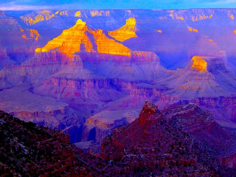 View of a canyon extending to the horizon. The canyon is shaded with sunlight reaching the peaks of the mountains within the canyon. In the foreground is the shaded edge of a cliff. The sky is blue with some white clouds on the horizon.