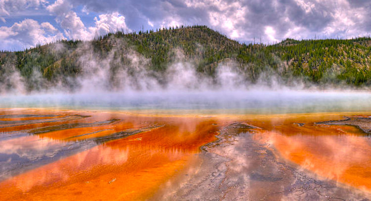 A volcanically active body of water with steam rising above. The water in the foreground appears orange or rust colored. The water further away appears to be blue. In the background are hills with green and brown foliage. The sky conditions are mostly cloudy, with sections of blue peeking through.