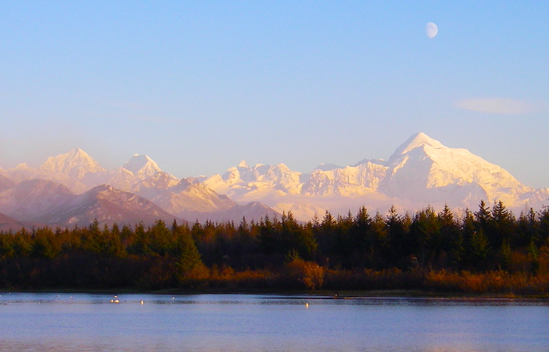 In the foreground is a lake with calm blue waters. The shore of the lake is forested, mostly with evergreen trees. In the background are tall mountains, the ones the furthest away are snow covered. The sky is blue with a three-quarter moon showing in the daylight.