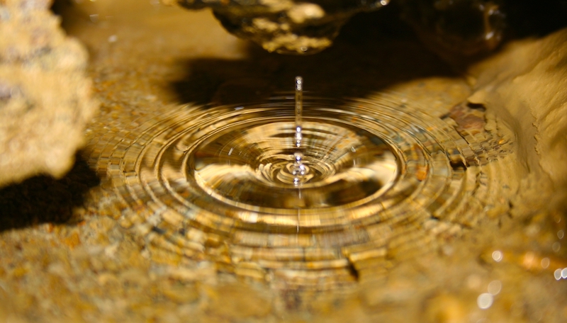 A clear pool of water inside a cave. A single drop disrupts the still surface, creating ripples. The bottom of the pool is covered in sediment and surrounded by brownish cave walls.