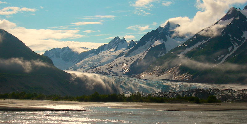 Glacier carving through valley to meet a body of water, surrounded by steep mountains with summer snow caps and evergreen trees along shore.